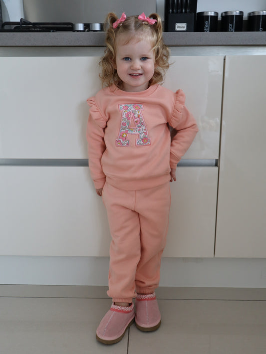 Child in pink tracksuit with a liberty floral initial A embroidered onto a pink frill sweater