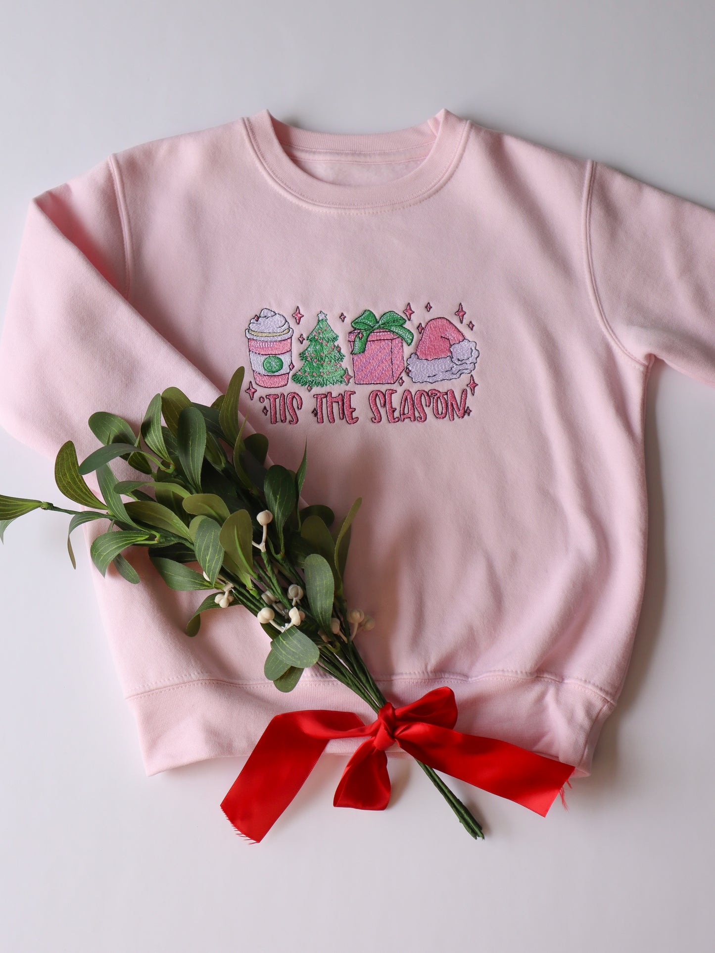 Tis the season kids pink embroidered sweater featuring a christmas design of a christmas tree, Latte, christmas gift and santa hat with the words tis the season