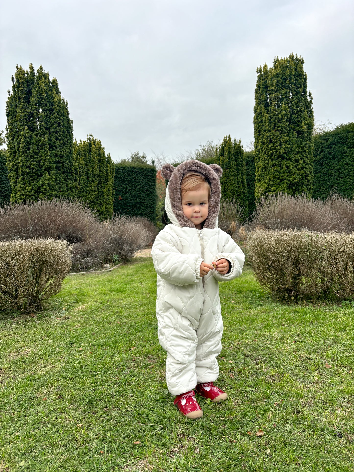 Child in a white snowsuit with bear ears standing in a garden.