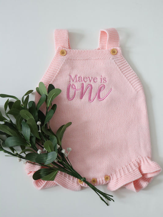 Pink knitted romper with 'Maeve is one' text on.