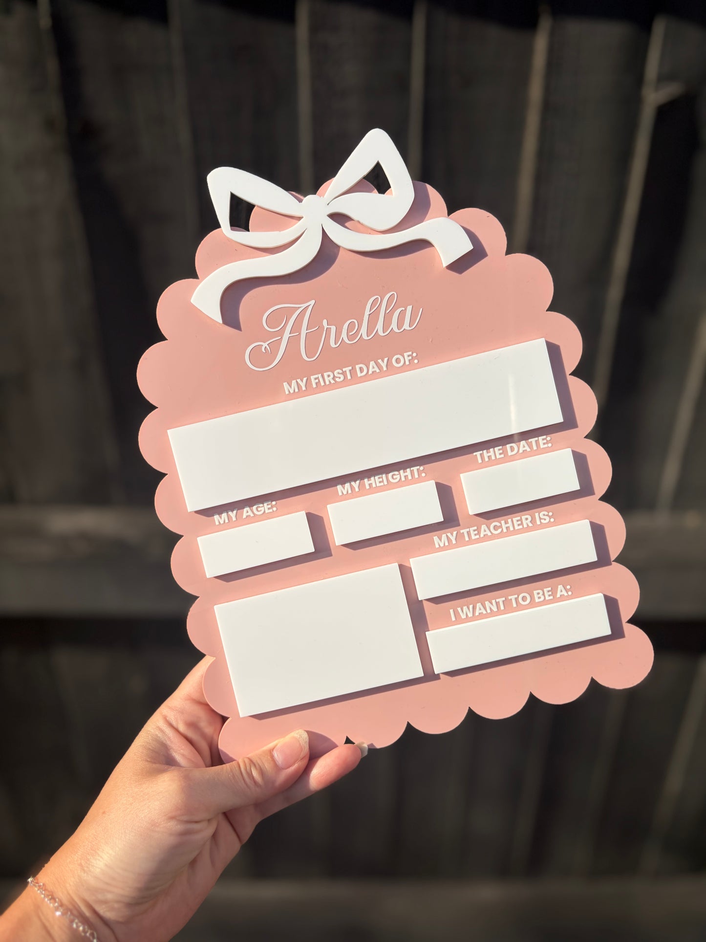 A hand holds a pink scalloped acrylic sign with a white bow at the top and labeled blank spaces for age, height, date, teacher, and aspirations.