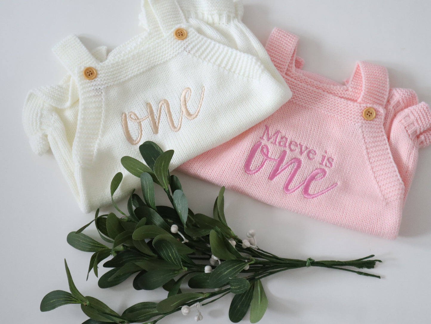Two baby outfits, one white and one pink, with embroidered text on a light background.