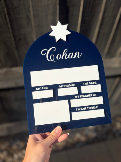 A hand holds a navy blue arched plaque or sign with a white star at the top and the name 'Cohan' in white script. The sign has blank white boxes for age, height, date, teacher, and other text, photographed against a blurred wooden fence background.