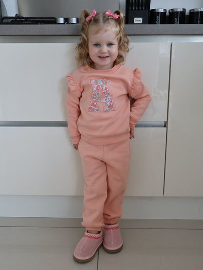 Child in pink tracksuit with a liberty floral initial A embroidered onto a pink frill sweater 