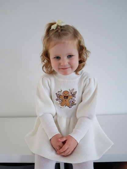 Young girl wearing a white knit sweater dress with a gingerbread man design against a plain background