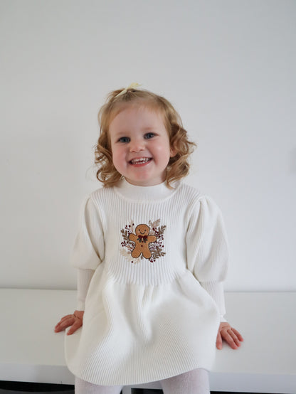 Child wearing a white sweater dress with a gingerbread man design on a plain background