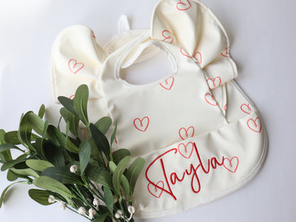 A white waterproof wing bib with red patterned hearts and the name 'Taylor' printed in red. The bib is displayed next to some green foliage.