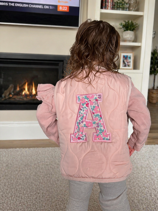 Child wearing a pink jacket with a floral letter 'A' in a living room.