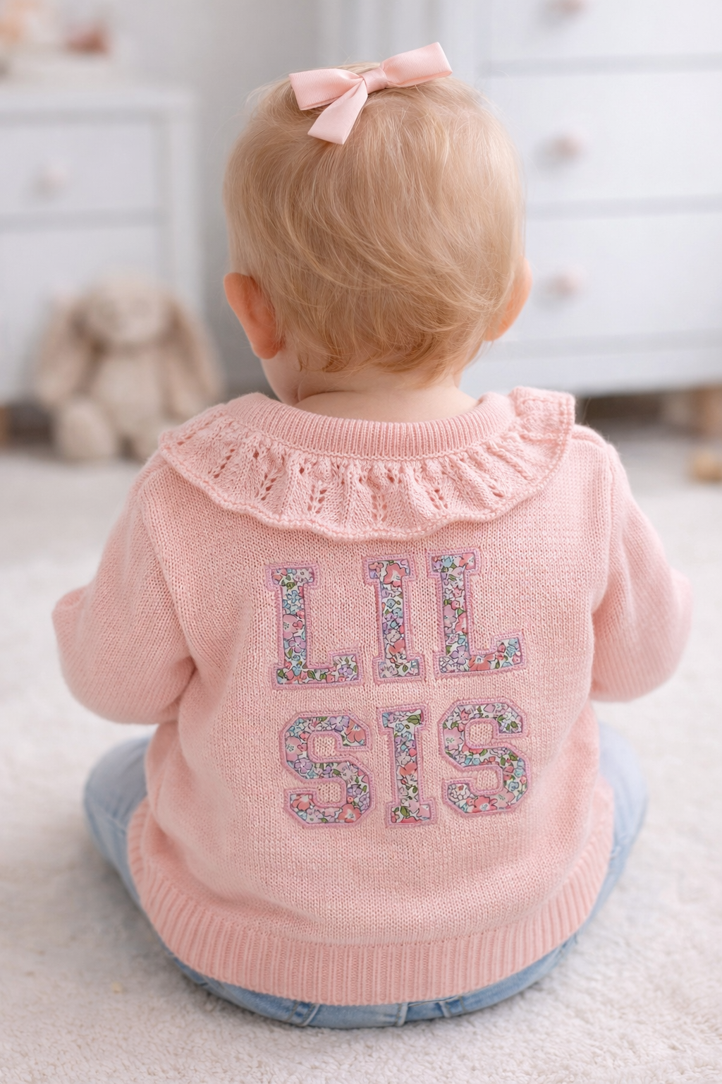 Baby wearing a pink cardigan with 'LIL SIS' text, standing in a room.