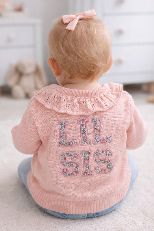 Baby wearing a pink cardigan with 'LIL SIS' text, standing in a room.