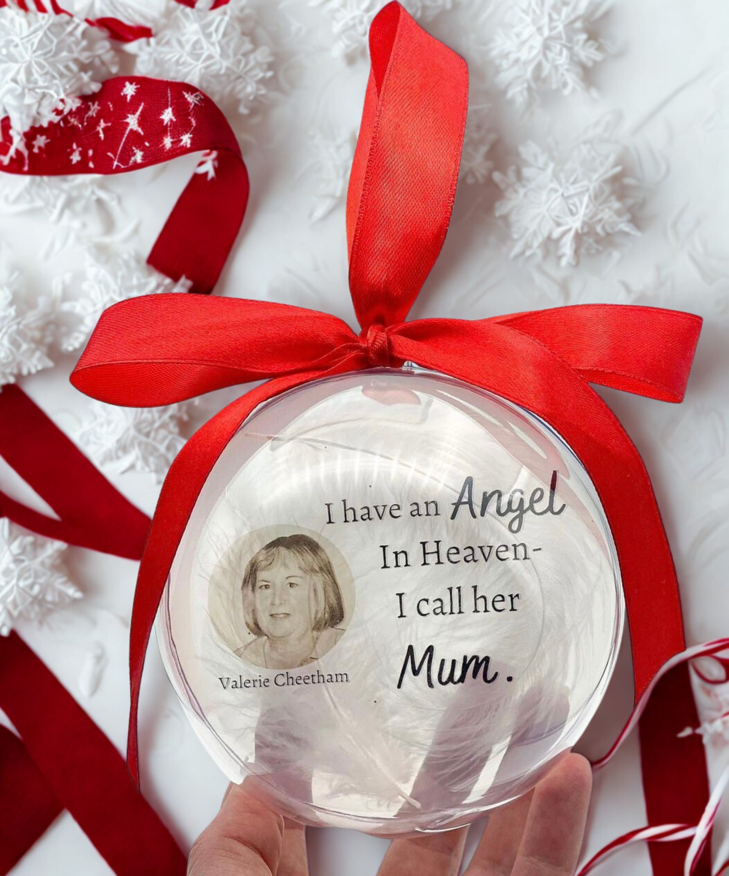 Decorative ornament with a red ribbon and text about an angel in heaven, held by a hand.