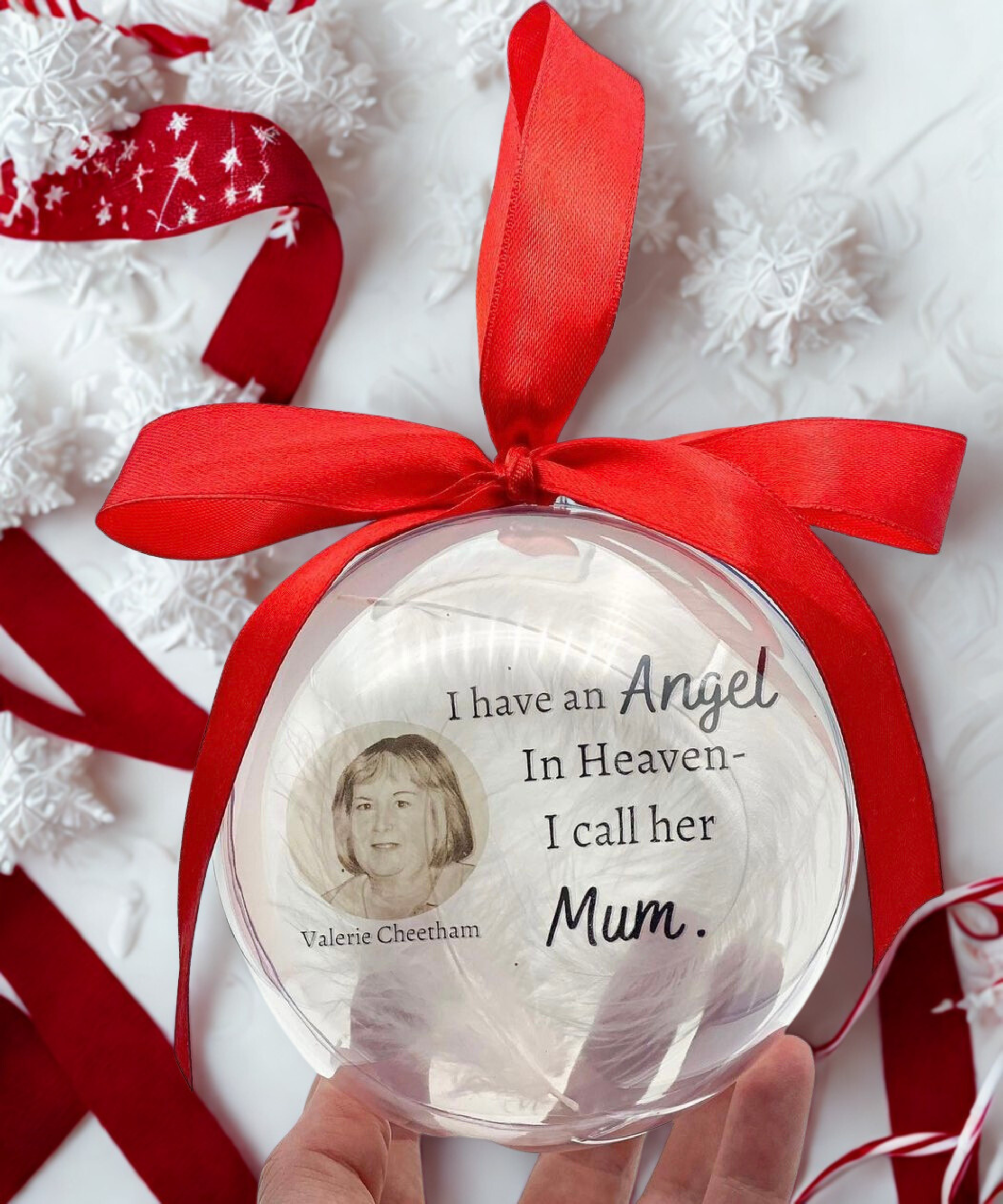 Decorative ornament with a red ribbon and text about an angel in heaven, held by a hand.