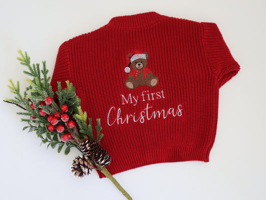 Red baby sweater with teddy bear design and 'My first Christmas' text, accompanied by festive decorations on a white background.