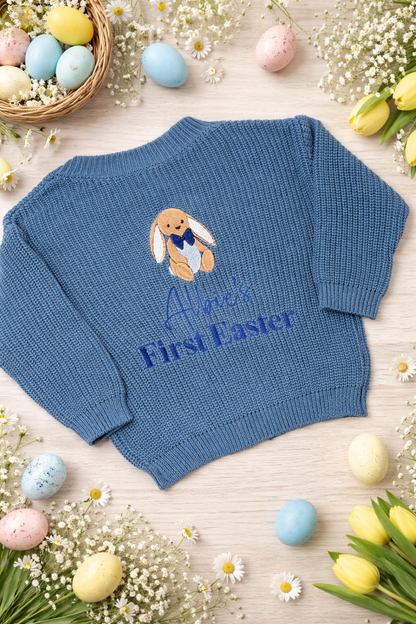 Blue baby sweater with embroidered bunny and 'Albie's First Easter' text, surrounded by Easter eggs and flowers on a light wooden surface.