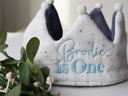 A fabric birthday crown with personalised name is One embroidered on it, in a color pattern with blue accents on the brim and a white fabric background.