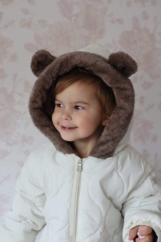Child wearing a white coat with a brown bear hood against a pink floral background