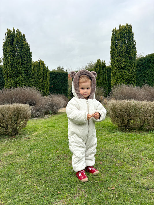 Child in a white snowsuit with bear ears standing in a garden.