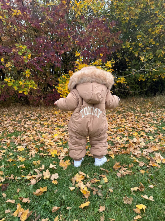Child wearing a brown snowsuit with a fur hood in an outdoor setting with trees and fallen leaves.