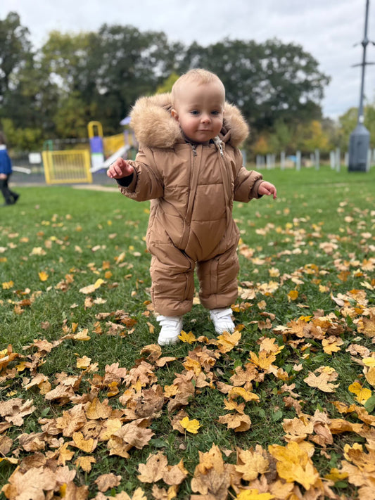 Baby in a brown snowsuit standing on grass with fallen leaves