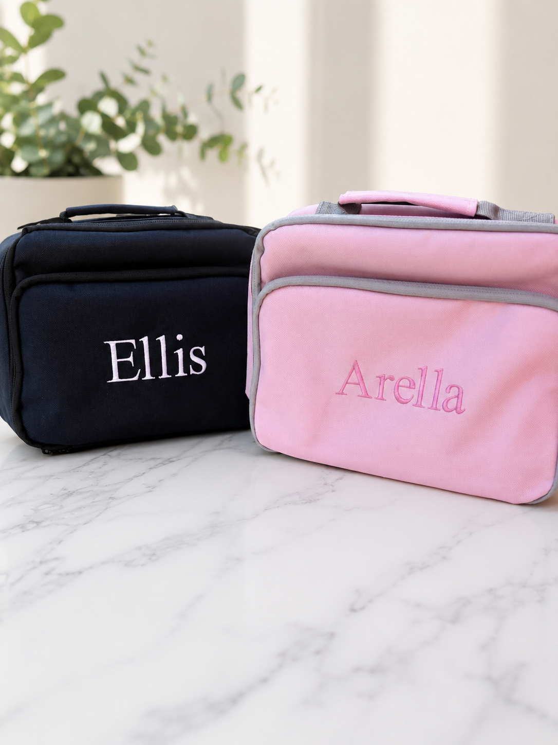 Two personalised soft-sided zippered childrens lunch bag on a white marble surface, one black with the name 'Ellis' in white and one pink with the name 'Arella' in pink