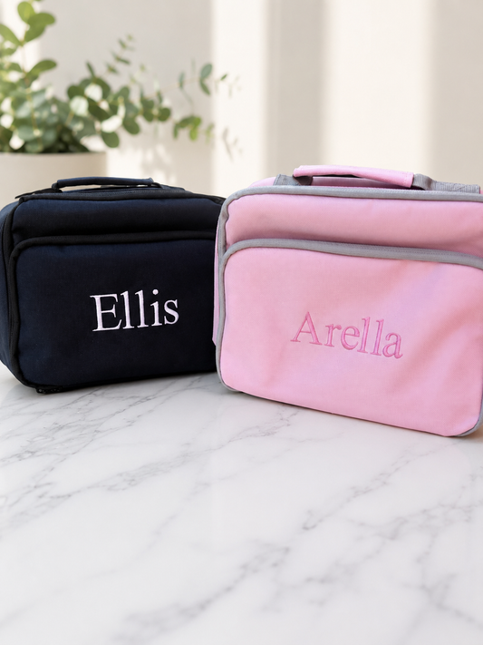 Two personalised soft-sided zippered childrens lunch bag on a white marble surface, one black with the name 'Ellis' in white and one pink with the name 'Arella' in pink