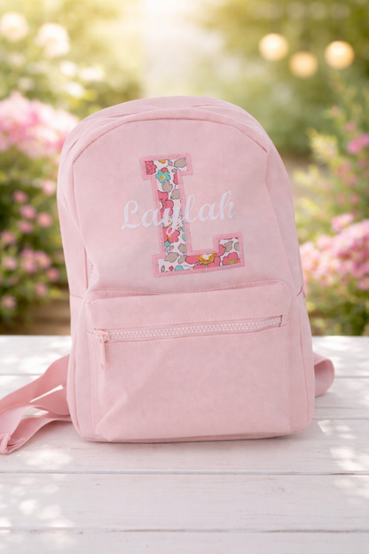 Pink backpack with floral letter 'L'  with the name Laylah embroidered through the middle on a wooden surface with blurred flowers in the background