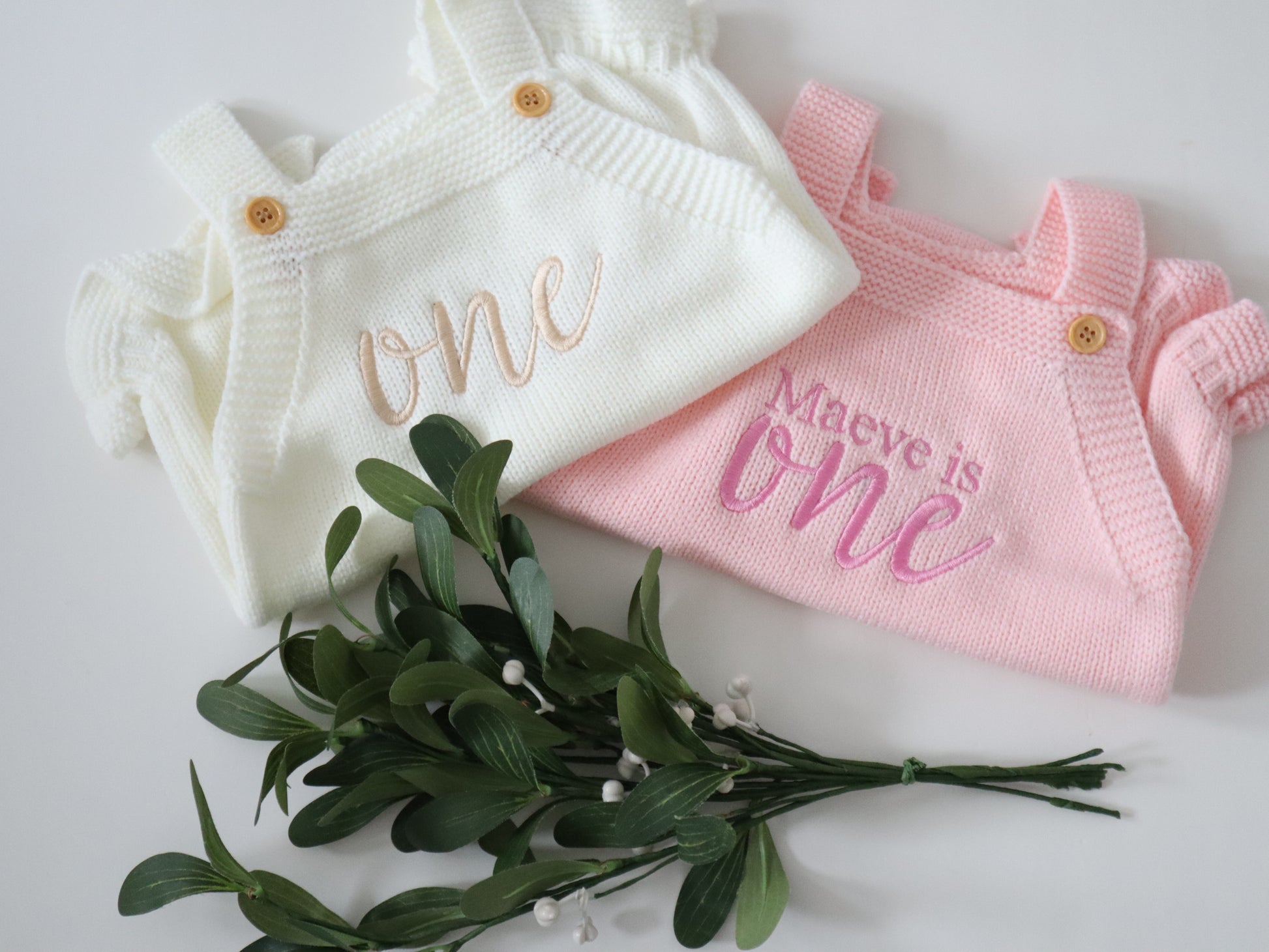 Two baby outfits, one white and one pink, with embroidered text on a light background.
