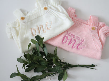 Two baby outfits, one white and one pink, with embroidered text on a light background.